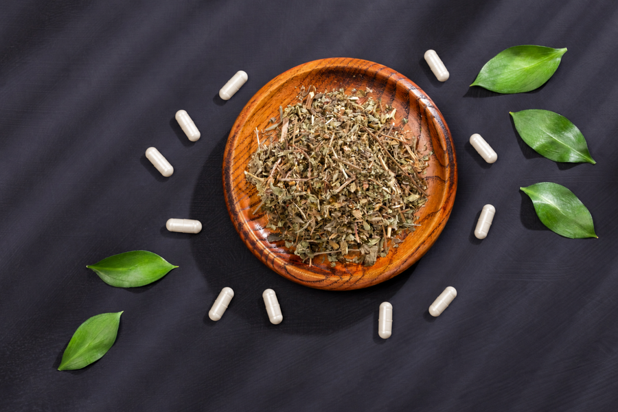 Wooden bowl of dried herbs surrounded by white supplement capsules and green leaves.