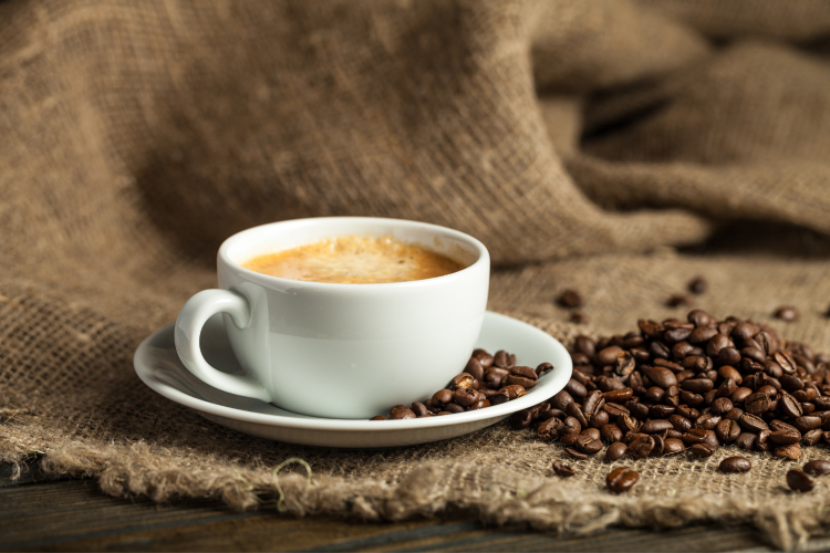 White coffee cup with crema next to whole roasted coffee beans on a burlap sack, illustrating the main subject of the kidney stones article.