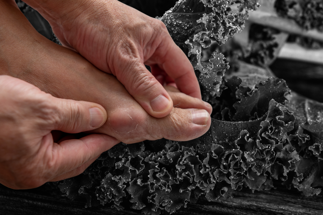Person holding their big toe during a gout flare-up, with kale in the background to suggest dietary support