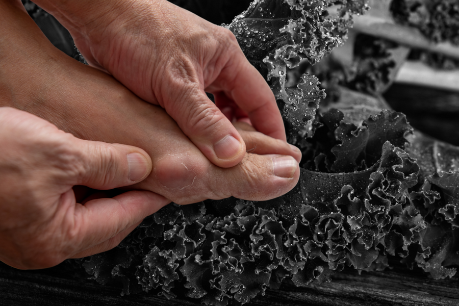Person holding their big toe during a gout flare-up, with kale in the background to suggest dietary support