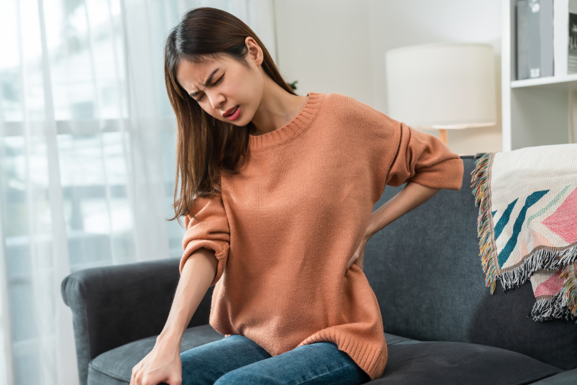 A person in an orange sweater and jeans sits on a couch, grimacing and holding their back.