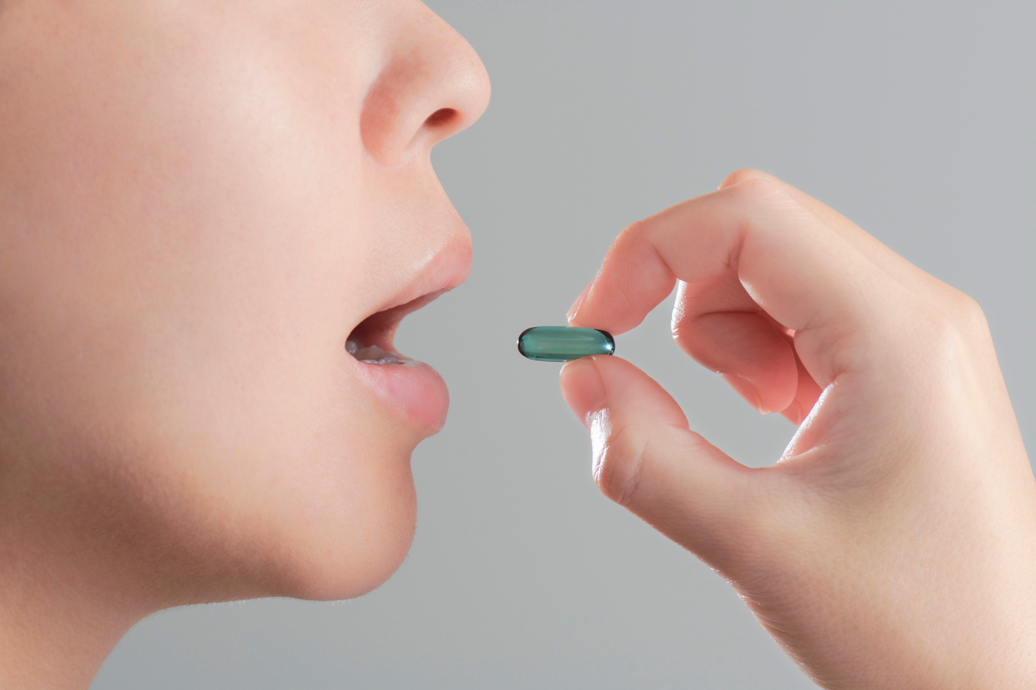 A hand holds a green capsule near an open mouth.