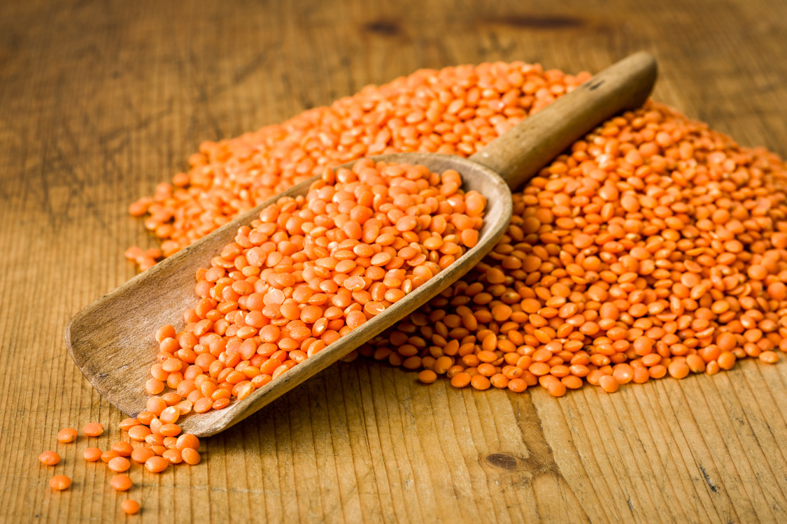 A wooden scoop piled with dry red lentils on a wooden table, illustrating the moderate purine content of lentils for gout management.