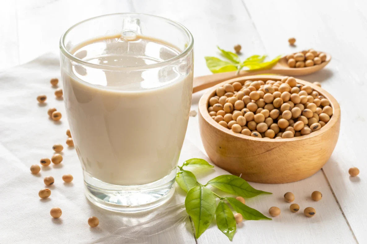 A glass of soy milk sits next to a wooden bowl and spoon filled with soybeans.