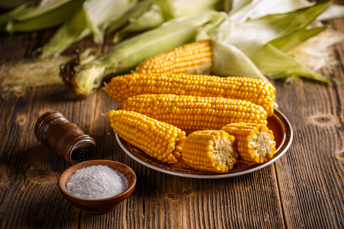 A plate of boiled or steamed corn on the cob, both whole and cut, with a small bowl of salt nearby, showing a gout-friendly preparation option for corn.