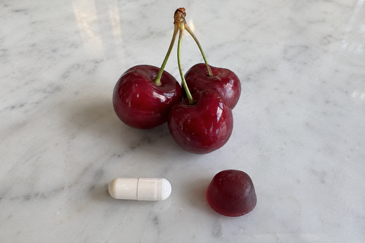 Three red tart cherries on a light marble surface, with a white capsule and a dark red gummy beneath them.