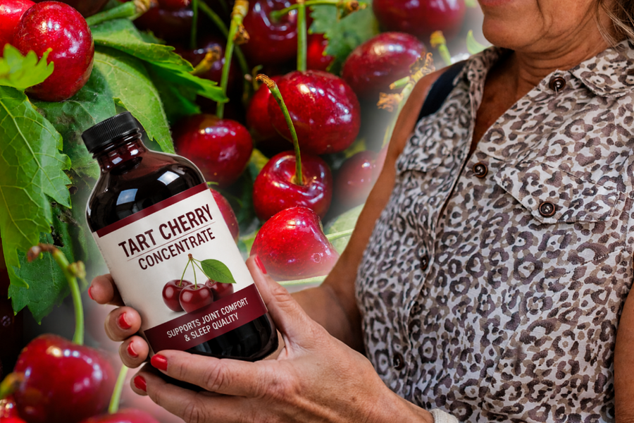 A person holds a dark bottle labeled “TART CHERRY CONCENTRATE” among fresh red cherries and green leaves, with the person’s patterned top visible in the background.