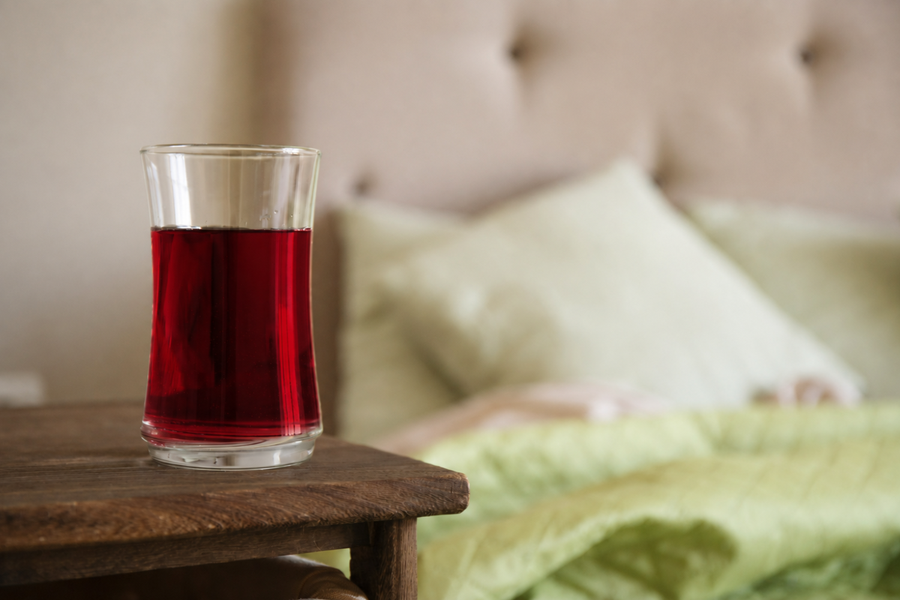 A clear glass filled with tart cherry juice sits on a wooden table, with a softly blurred bed and green-and-white bedding in the background.