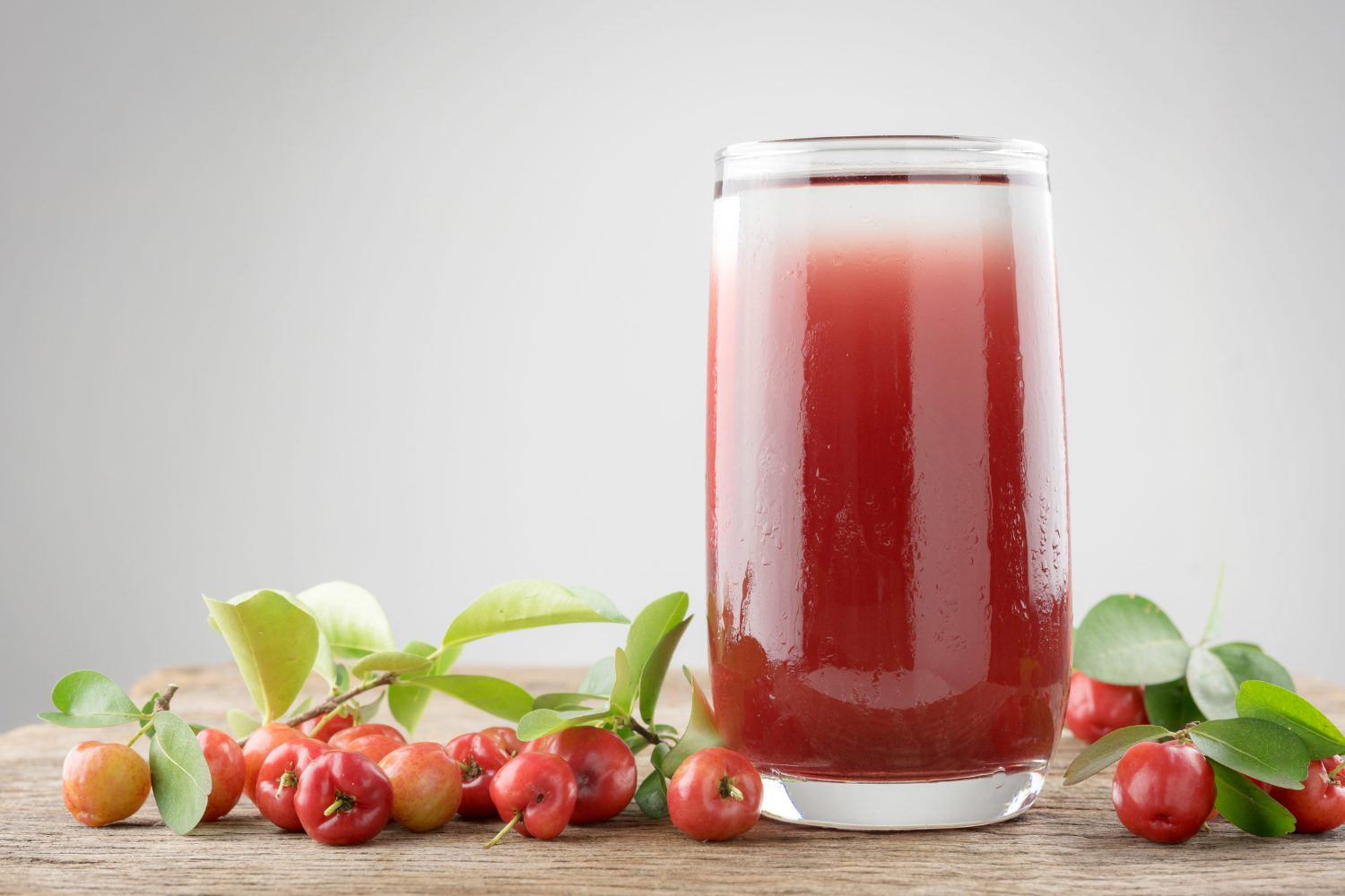 A glass of red juice sits on a wooden table, surrounded by acerola cherries and green leaves.