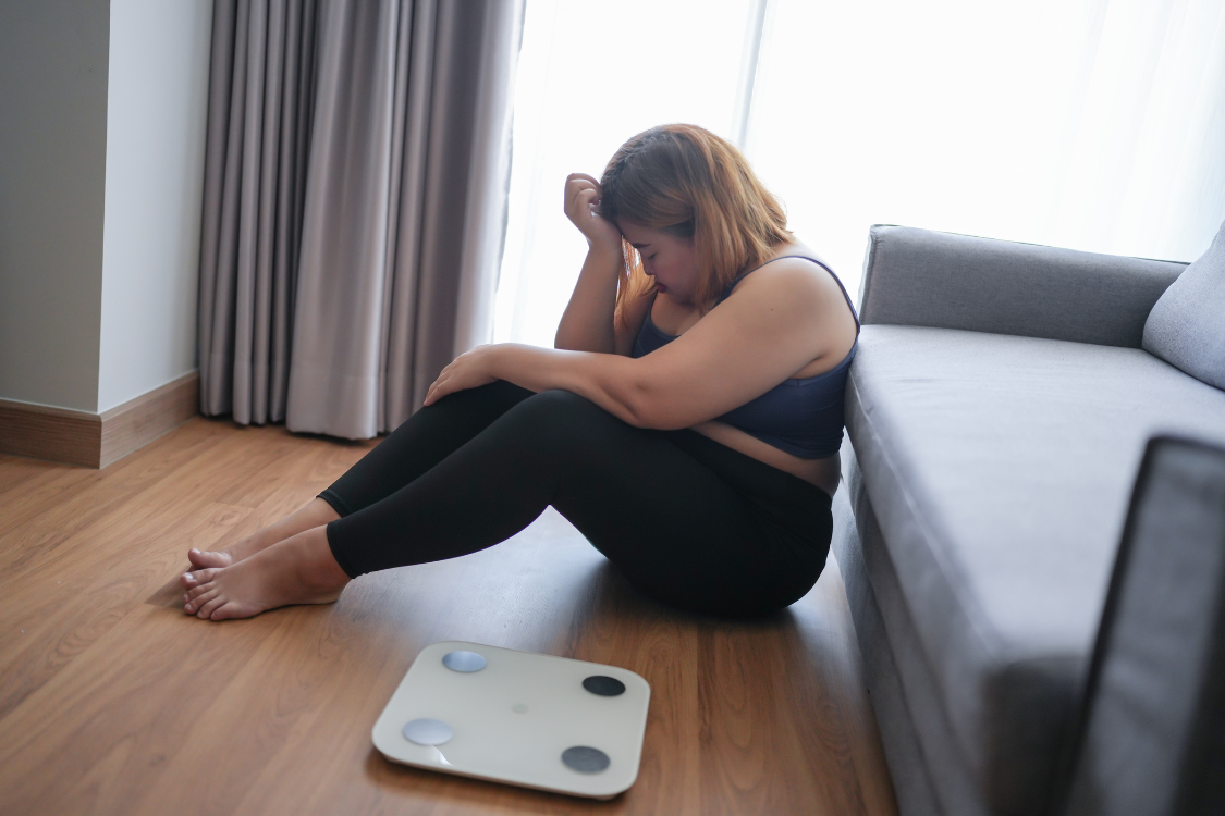 Overweight woman sitting on floor looking discouraged next to a weight scale, struggling with obesity and gout risk